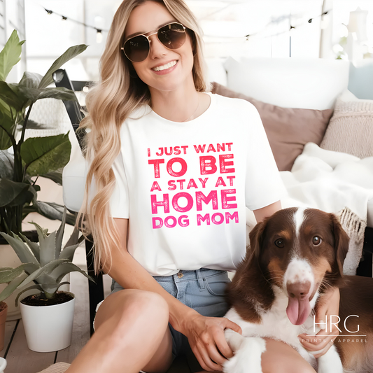 Woman wearing a "Stay At Home Dog Mom" graphic tee with bold pink text, sitting with her dog, surrounded by plants and cozy decor.